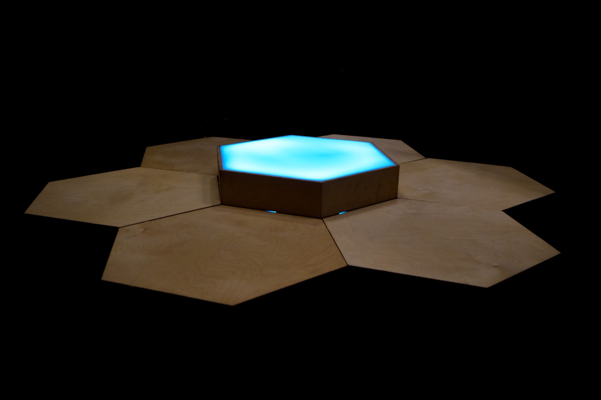 Resident Thrum: the hexagonal grid of seven plywood tiles with the central acrylic tile glowing blue, on a black floor